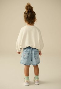 Maglione bianco texturizzato, pantaloni shorts in denim blu con ricami floreali decorativi sulle tasche e sneakers colorate. Bambino di spalle rispetto alla fotocamera.