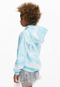 Lichtblauwe tie-dye hoodie met een voorzak en capuchon, gecombineerd met een korte, sprankelende tulle rok, die doorschijnende lagen en patronen laat zien.