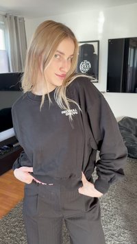 Mladá žena s dlhými blond vlasmi, nosiaca čiernu mikinu Red Bull Racing a čierne nohavice, pózujúca v modernom obývacom izbe.