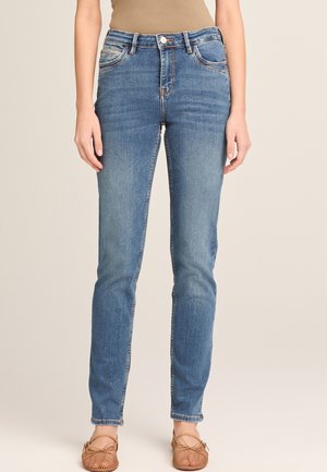 Schmal geschnittene blaue Jeans, getragen von einer stehenden Person, kombiniert mit hellbraunen, gewebten Ballerinas vor neutralem Hintergrund.