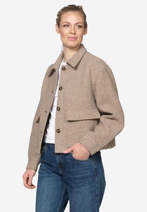 Woman wearing beige buttoned wool jacket over white top and blue jeans, standing with one hand in pocket, looking to the side.