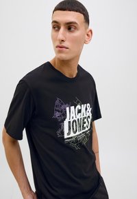 Sort bomulds T-shirt med et hvidt og farverigt grafisk design med teksten "JACK & JOVES." Rund hals, korte ærmer, afslappet pasform.