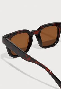 Lunettes de soleil en écaille noire et brune avec des verres carrés et teintés marron, dotées d'une monture en plastique épaisse et glossy et de branches fines.