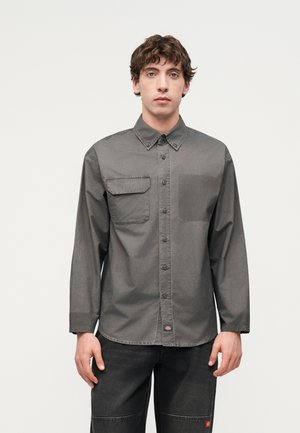 Jeune homme portant une chemise gris foncé boutonnée avec une poche poitrine et un jean noir, debout devant un fond blanc uni.