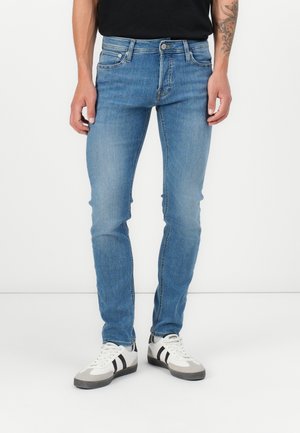 Homme portant un jean slim bleu et des baskets blanches avec des rayures noires, debout sur un fond blanc uni.