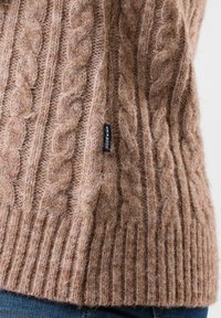 Brązowy sweter w splot kablowy z prążkowanym wykończeniem i miękką teksturą. Mała metka z boku z nazwą marki. Zaprojektowany z myślą o ciepłu i komforcie.
