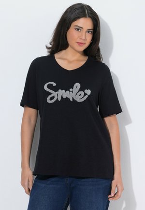 Ung kvinde med mørkt hår iført en sort t-shirt med teksten "Smile" og et hjerte i tekstureret grå skrift, kombineret med blå jeans.
