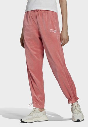 Personne portant un pantalon de jogging en velours rose avec un cœur et le logo Adidas sur la cuisse, ainsi que des baskets blanches épaisses sur un fond uni.
