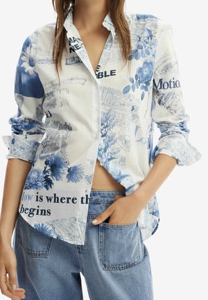 Frau trägt ein blau-weißes Hemd mit Blumen- und Schriftmuster, dessen Ärmel hochgekrempelt sind, kombiniert mit hoch taillierten blauen Jeans.