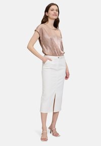 Ein rosegoldenes, geripptes Top, kombiniert mit einem weißen Denim-Midirock, das mit einem Schlitz vorne und Taschen ausgestattet ist. Das Model trägt neutrale Sandalen.