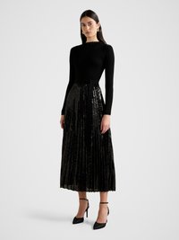 Black ribbed long-sleeve top paired with a pleated black sequined skirt, accented by a tied waistband, complemented by black stiletto heels.