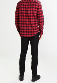 Mężczyzna w czerwono-czarną kratkę z długim rękawem, czarnych spodniach i czarnych butach, stojący tyłem na jednolitym tle.