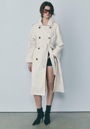 Donna che indossa un trench doppiopetto color avorio con cintura, mini gonna nera, stivaletti con tacco neri e grandi occhiali da sole, in piedi contro una parete liscia.