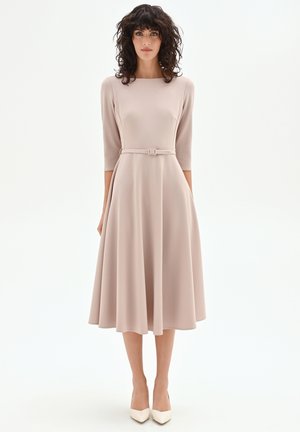 Femme aux cheveux bouclés foncés portant une robe midi beige à manches trois-quarts, ceinturée, et des escarpins blancs à bout pointu, debout devant un fond blanc.