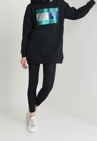 Czarna, oversize bluza z kapturem z teksturowaną, wielokolorową łatą, w zestawie z czarnymi legginsami i białymi trampkami. Gładki materiał i swobodny krój.