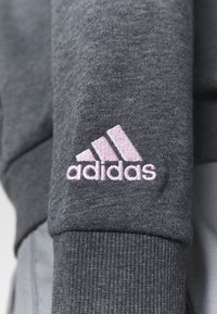 Ciemnoszary materiał sweatshirtu z ribbowanym mankietem i różowym haftowanym logo Adidas z trzema ukośnymi paskami.