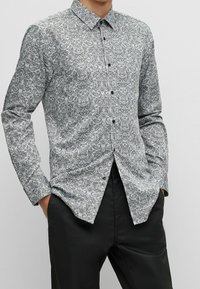 Camisa de manga larga con botones, en patrón paisley blanco y negro, con cuello clásico y botones negros, combinada con pantalones oscuros.