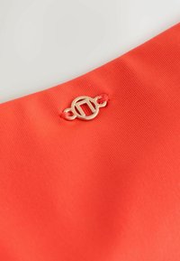 Petit fermoir circulaire doré avec un motif découpé attaché à un tissu côtelé orange vif.