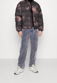 Hombre con una chaqueta de plumas de tie-dye en negro y rosa, pantalones de pana morados y zapatillas blancas, de pie contra una pared clara.