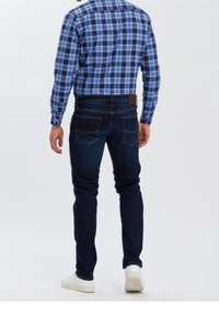 Muž v tmavě modrých džínách, modré kostkované košili a bílých teniskách, stojící proti bílému pozadí, zády k divákovi.