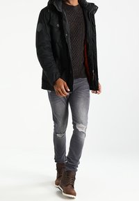 Mann trägt eine schwarze Jacke, einen dunkelbraunen Strickpullover, zerrissene graue Jeans und braune Schnürstiefel und geht vorwärts.