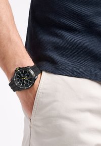 Reloj negro con una correa de goma texturizada, cara redonda con marcas en blanco y subesferas, llevado en una muñeca, junto a pantalones beige.
