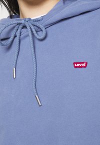 Detailní záběr modré mikiny s kapucí, šňůrky uvázané do mašle a malým červeným logem Levi's na hrudi.