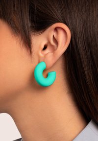 Orecchino a cerchio di un bel turchese opaco, con un'apertura parziale. Design leggero con una texture liscia, indossato all'orecchio di un modello.