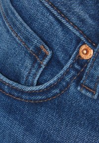 Primer plano de tela de mezclilla azul que muestra costuras dobles en marrón, el borde del bolsillo y un remache de cobre en los vaqueros.