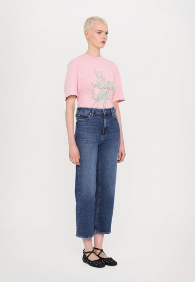 Pink cotton t-shirt with green text, paired with blue wide-leg jeans and black polka dot flats with crossed straps.