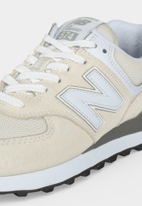 Μπεζ και λευκό αθλητικό παπούτσι New Balance 574 Classic με λευκά κορδόνια, επάνω μέρος από δίχτυ και σουέτ και μαύρη υφασμένη σόλα.