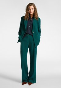 Una donna sta in piedi con le mani nelle tasche, indossando un completo turchese, una camicia abbottonata blu navy e scarpe a punta marroni su uno sfondo chiaro e semplice.