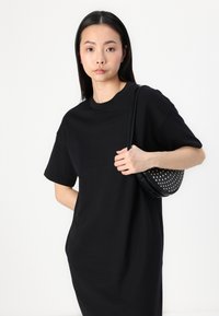 Černé oversized tričko s krátkým rukávem a kulatým výstřihem. Modelka drží texturovanou černou tašku s perforovanými detaily.