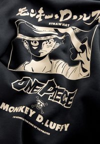 Sort stof med et grafisk tryk af en figur med en stråhat, omgivet af en cremefarvet kontur. Teksten inkluderer "One Piece" og "Monkey D. Luffy."