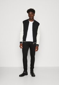 Chaqueta bomber negra con mangas blancas, pantalones ajustados negros y botas negras hasta el tobillo. La chaqueta cuenta con un cuello acanalado y un detalle minimalista.