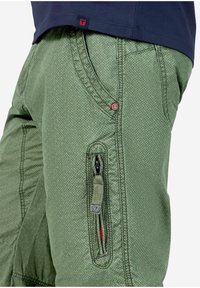 Grüne, strukturierte Hosen mit einem Rautenmuster, ausgestattet mit schrägen Vorderaschen und einer seitlichen Ziptasche mit roten Akzenten und einem Logo-Button.