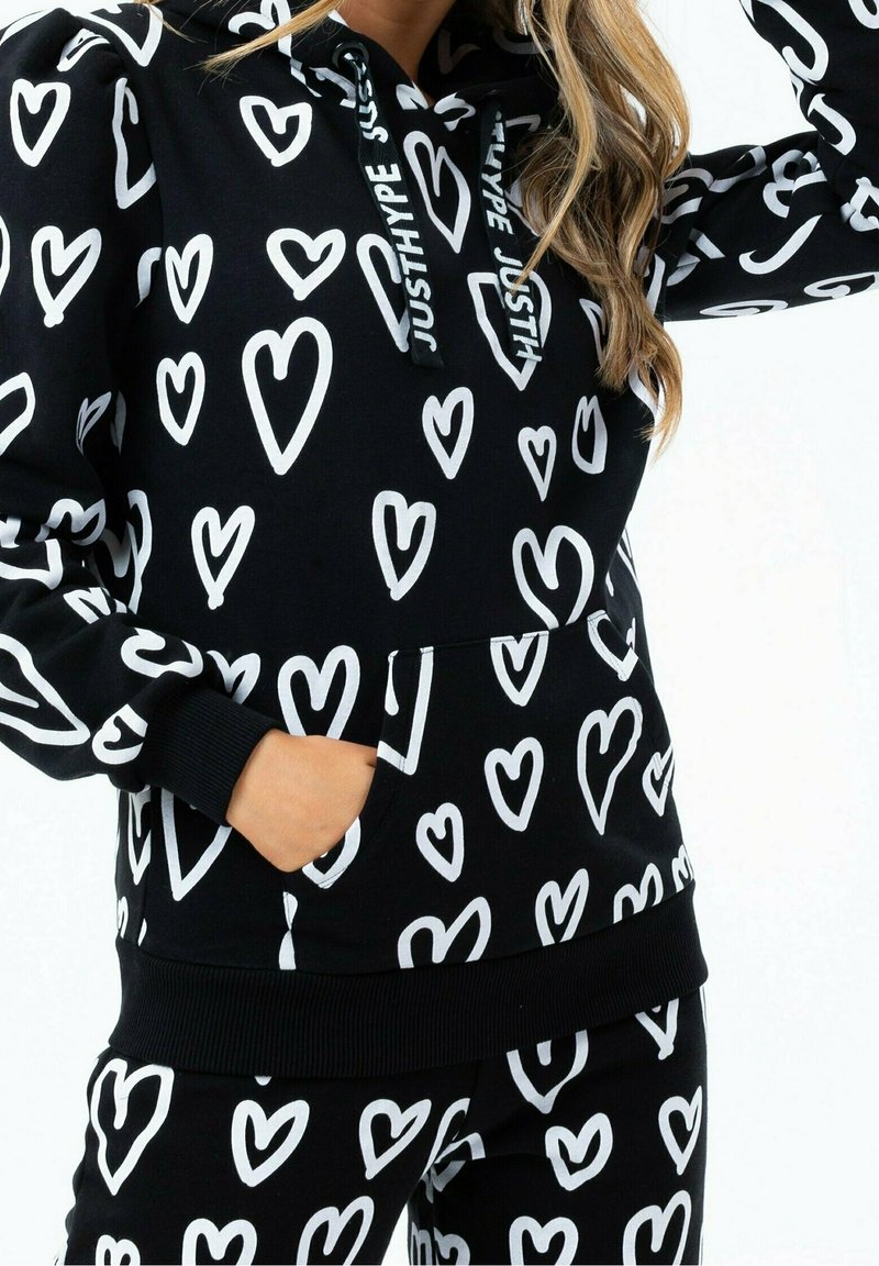 Femme portant un sweat à capuche noir et un pantalon avec des motifs de cœurs blancs, cordons du sweat portant l'inscription "JUSTHYPE,", main dans la poche avant.