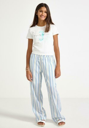 Hymyilevä tyttö seisoo valkoisessa T-paidassa, jossa on sininen merihevosen kuvio, väljissä valkoisissa housuissa, joissa on siniset raidat, valkoisissa sandaaleissa ja rannekoruissa.