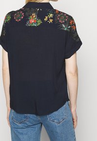 Camicia blu navy con yoke a fantasia floreale. Maniche corte, vestibilità rilassata e tessuto morbido. Orlo posteriore leggermente arrotondato.