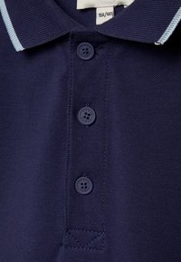 Polo shirt blu navy con trama a maglia, caratterizzato da una chiusura con tre bottoni e una striscia blu chiaro sul collo. Bottone con dettaglio del logo in rilievo.
