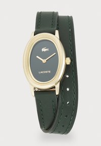 Montre ovale dorée avec bracelet en cuir vert foncé, présentant une lunette texturée, le logo Lacoste et des aiguilles des heures et des minutes dorées.