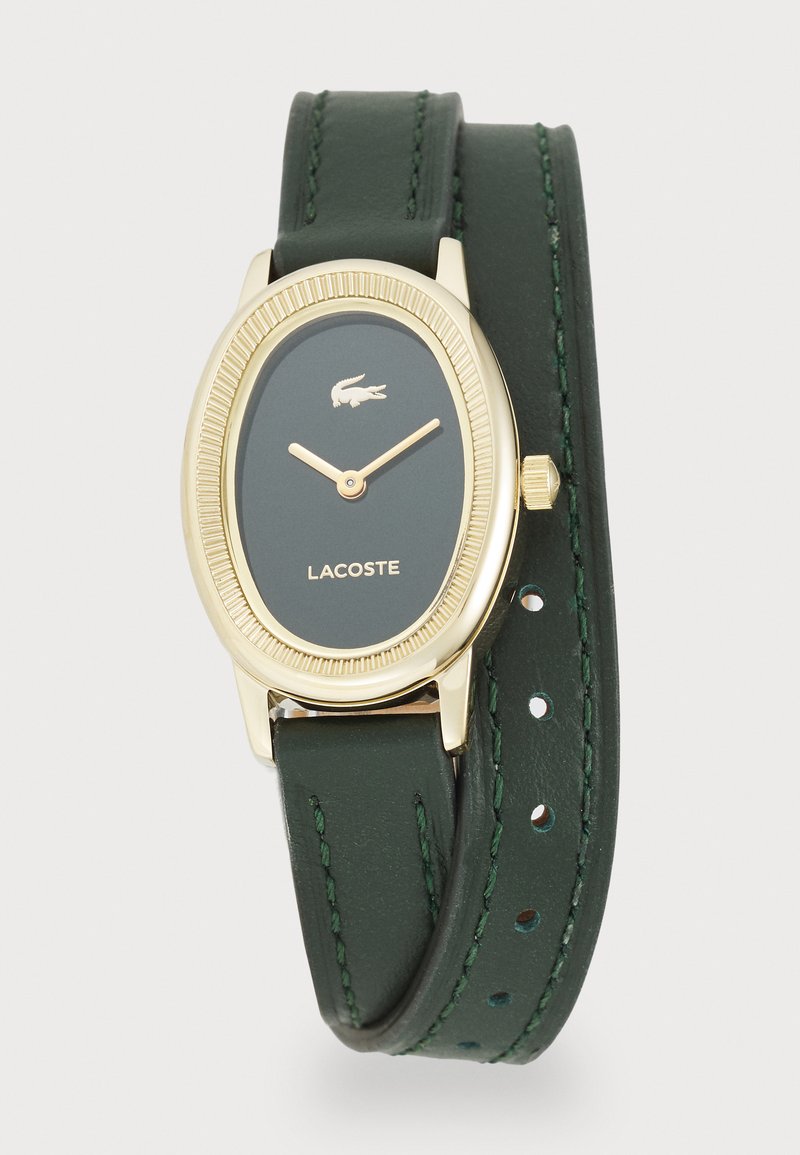 Montre ovale dorée avec bracelet en cuir vert foncé, présentant une lunette texturée, le logo Lacoste et des aiguilles des heures et des minutes dorées.