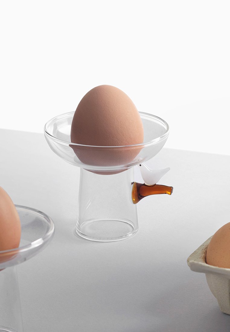Przezroczysty szklany uchwyt na jajka z zaokrąglonym talerzykiem, ozdobiony stylizowanym uchwytem w kształcie ptaka w białym i pomarańczowym kolorze, podtrzymujący jasnobrązowe jajko.