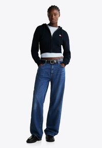 Geplukt zwart kabelgebreid hoodie met rits, lichtblauw T-shirt eronder, wijdvallende blauwe jeans, zwarte riem en zwarte loafers met textuurdetail.