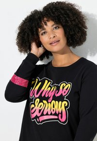 Maglietta nera a maniche lunghe con una grafica colorata "Why So Serious" sul davanti e un'accento di paillettes rosa sulla manica sinistra.