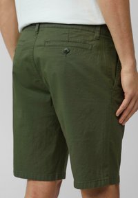 Shorts en coton verts à coupe décontractée, avec une poche arrière à bouton et des bords ourlés. Texture visible et détails de design simples.