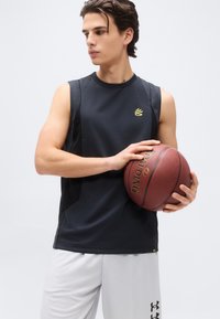 Νέος άνδρας με μαύρο αθλητικό μπλουζάκι χωρίς μανίκια και άσπρο σορτς κρατά μια μπάλα του μπάσκετ, κοιτάζοντας προς τα αριστερά σε απλό φόντο.