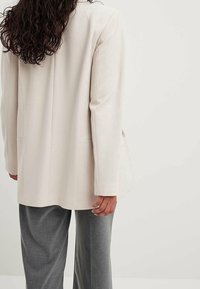 Světle béžový dlouhý blazer s hladkým materiálem, dvěma předními kapsami a lehce oversize střihem, spárovaný se šedými kalhotami se širokými nohavicemi.