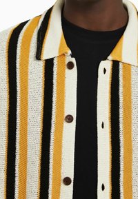 Sweter z dzianiny z poziomymi paskami w kolorze żółtym, czarnym i kremowym. Brązowe guziki oraz kołnierzyk. Miękki, teksturowany materiał.