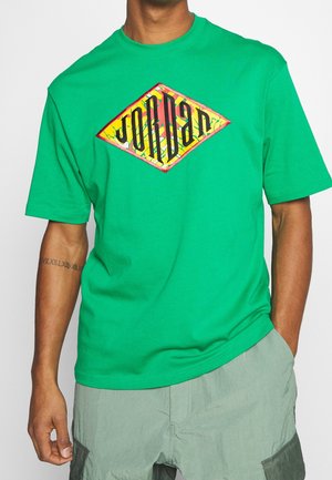 Zielona bawełniana koszulka z krótkim rękawem, ozdobiona kolorowym logo w kształcie diamentu z napisem 'Jordan' oraz luźnym krojem. Tło jest jasne, białe.